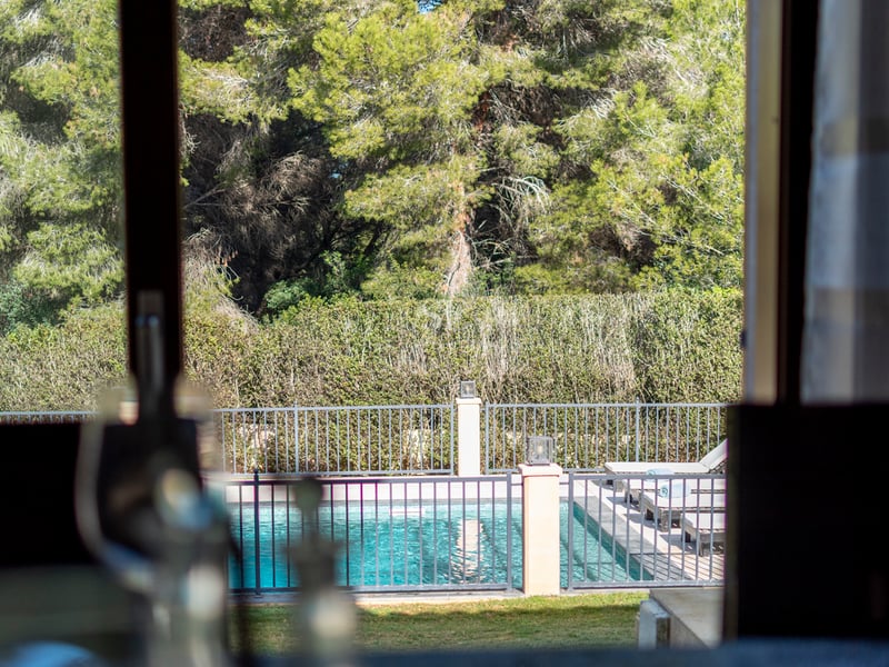 Utsikt genom ett fönster mot en inhägnad turkos pool omgiven av solstolar och höga tallar.