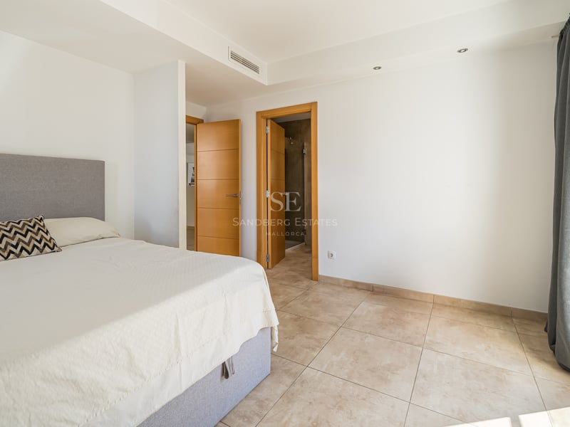 Ett soligt sovrum med grå säng, klinkergolv, vita väggar och trädörrar som leder till ett badrum.