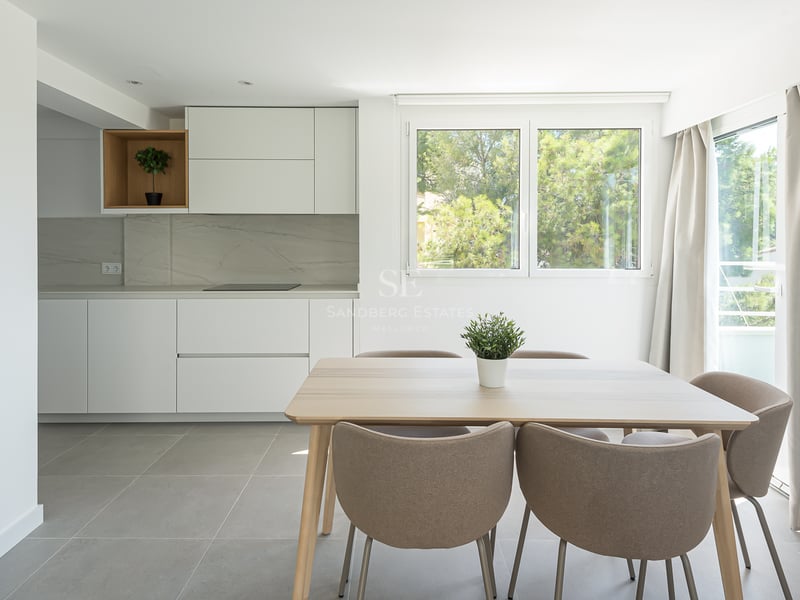 Salle à manger lumineuse avec table en bois clair, chaises taupe et cuisine blanche minimaliste vue sur jardin.