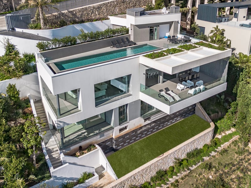 Vue aérienne d'une villa blanche à trois niveaux avec piscine à débordement sur le toit et balcons en verre.
