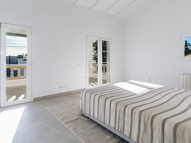 Minimalistiskt sovrum med randig säng, grått stengolv, högt tak med vita bjälkar och solig balkong.