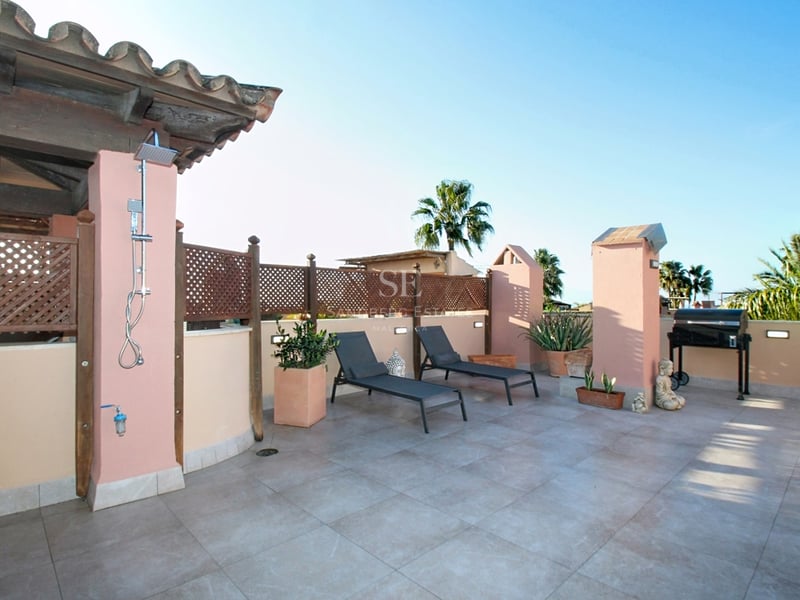 Stor medelhavsterrass med solstolar, utedusch, grill och ljusa stengolv under en klarblå himmel.