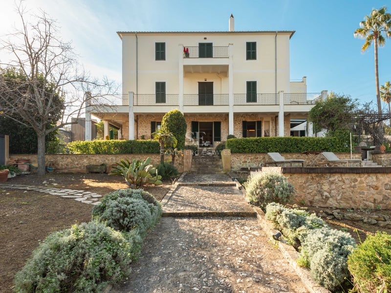 Vy av en trevåningsvilla med stendetaljer, gröna fönsterluckor och en stenstig genom en prunkande trädgård.