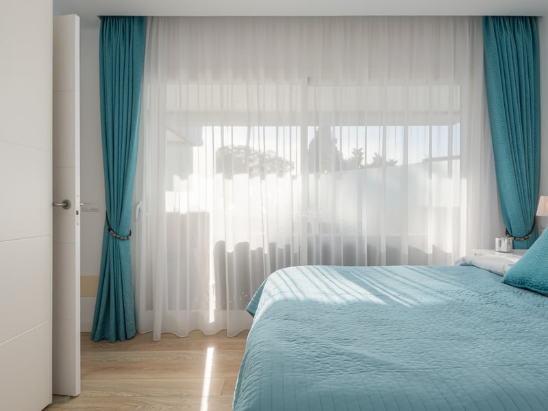 Chambre lumineuse avec lit turquoise, rideaux assortis et grandes fenêtres laissant entrer la lumière naturelle.