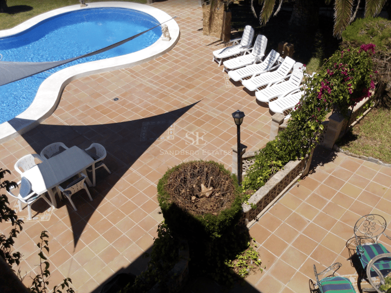 Vista aérea de una piscina azul en forma de riñón rodeada de baldosas de terracota, tumbonas blancas y zona de comedor.
