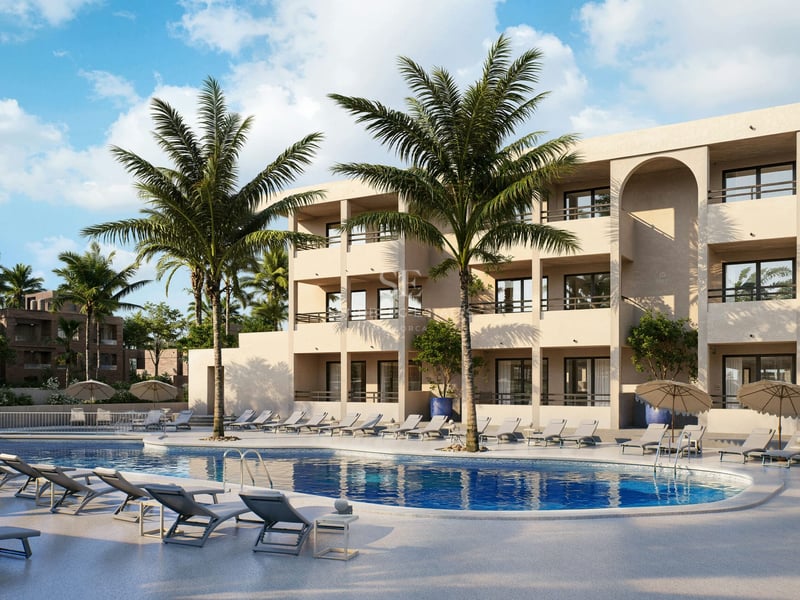 Grande piscine bleue avec chaises longues, palmiers et bâtiment méditerranéen moderne de trois étages sous un ciel clair.