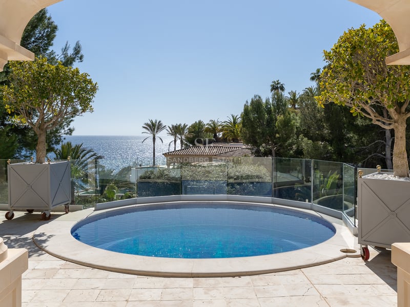 Runder blauer Pool auf einer Steinterrasse mit Glasgeländer und Blick auf das Mittelmeer und Palmen.