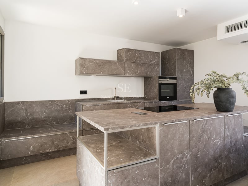 Modernt kök med stor köksö i gråmarmorerad sten, inbyggda vitvaror och minimalistiska vita väggar.