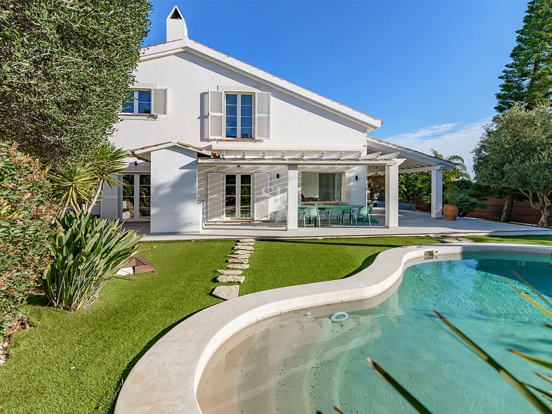 Baksida av en vit villa med en turkos pool, grön gräsmatta och en stenstig under en klarblå himmel.