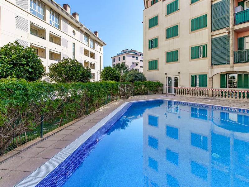 Utomhuspool med blå mosaik omgiven av stenplattor och byggnader med klassiska gröna fönsterluckor.