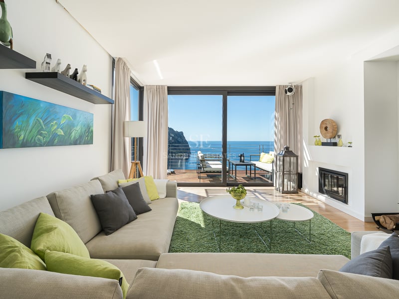 Bright modern living room with large glass doors opening to a sea-view terrace, featuring a neutral sectional sofa.