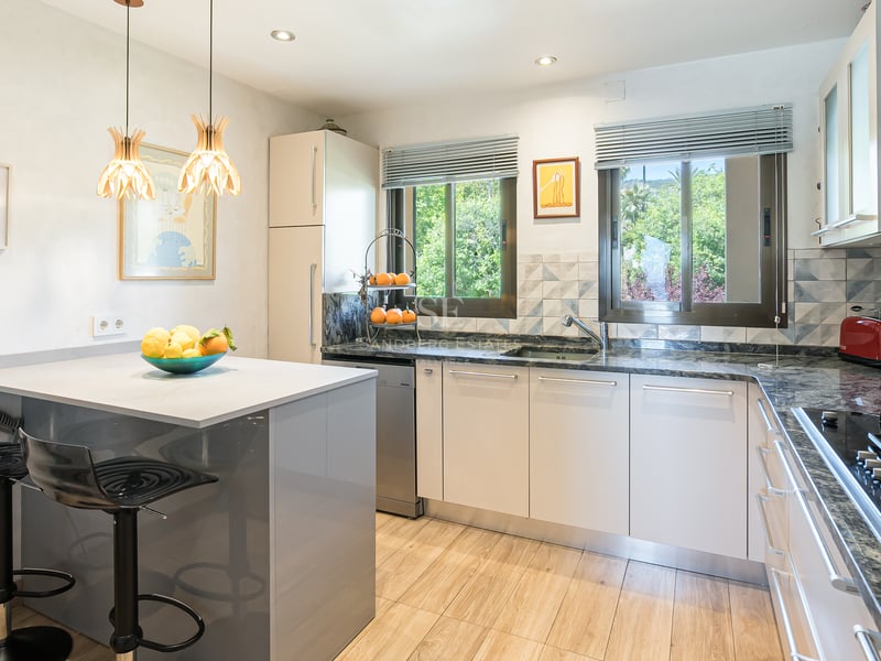 Modernt kök med vita skåp, bänkskivor i granit, köksö och fönster med utsikt över grönska.