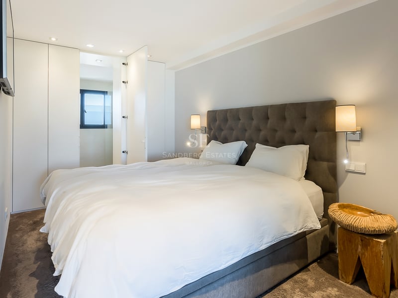 Modernes Schlafzimmer mit grauem gestepptem Kopfteil, weißen Einbauschränken und einem handgeschnitzten Holztisch.