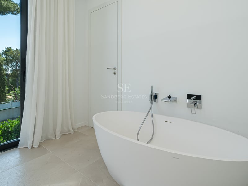 Baignoire îlot ovale blanche à côté d'une baie vitrée offrant une vue sur des pins méditerranéens luxuriants.