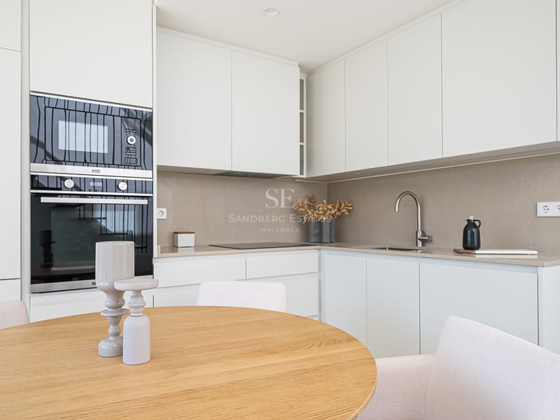 A bright minimalist kitchen featuring white cabinetry, sand-colored backsplash, integrated oven, and a wooden dining table.