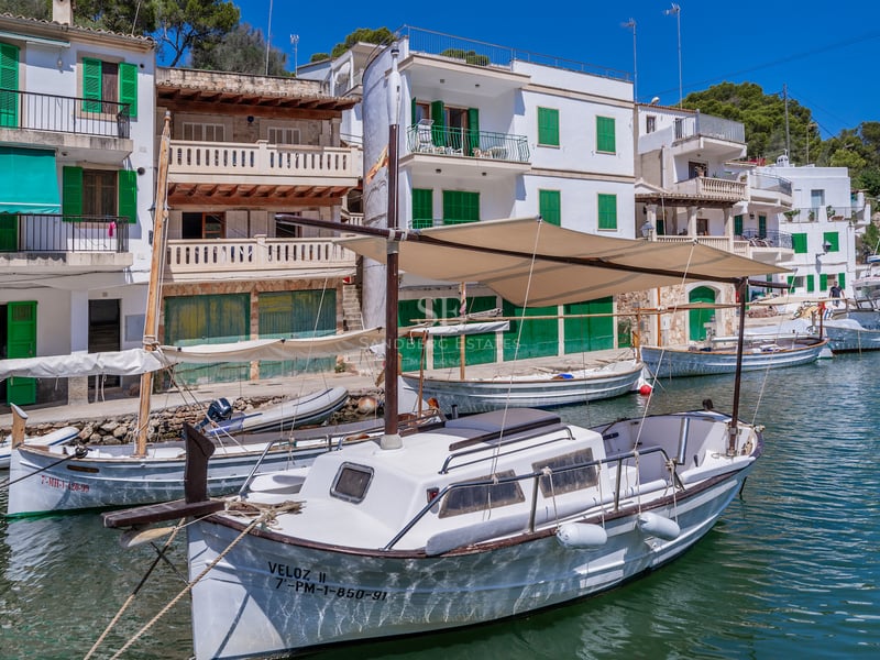 Maisons blanches aux volets verts surplombant un port paisible avec bateaux en bois et eau turquoise.
