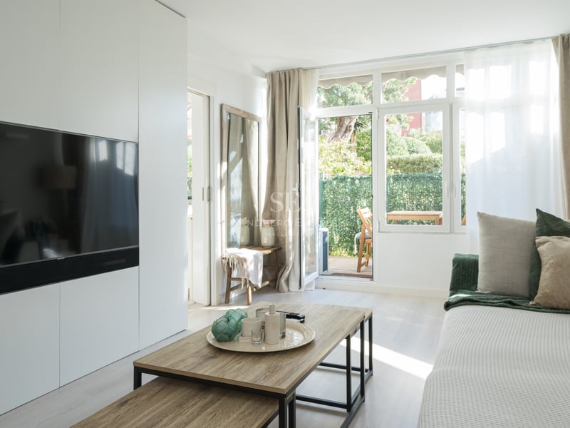 Modern living room with white wall unit, integrated TV, wooden coffee table, and glass door opening to a private garden.