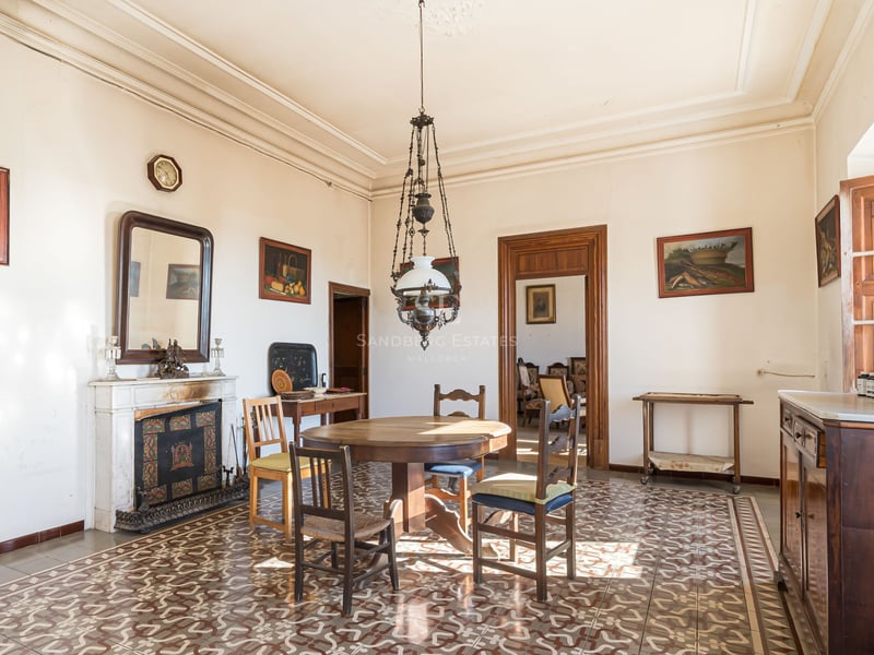 Salle à manger ancienne avec table en bois, sol en carrelage orné, cheminée en marbre et hauts plafonds.