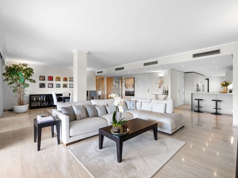 Spacieux salon avec canapé en L blanc, table basse en bois sombre, sol en marbre et cuisine intégrée.