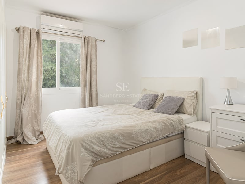 Chambre lumineuse avec lit double, meubles blancs, parquet et fenêtre donnant sur des arbres.