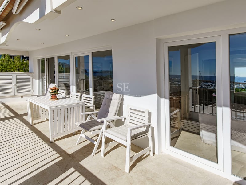 Sunlit terrace with white outdoor dining set and large glass sliding doors reflecting the coastline.