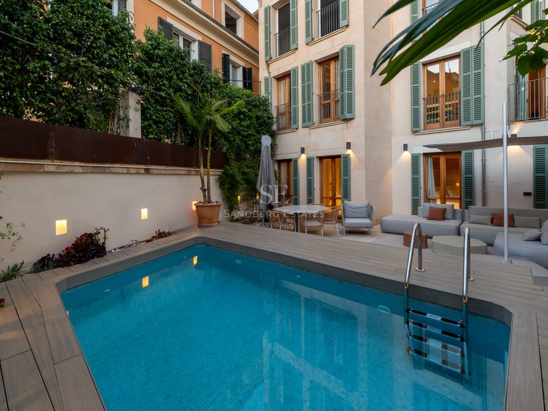 Piscine rectangulaire bleue à côté d'un salon moderne et d'un bâtiment traditionnel aux volets verts.