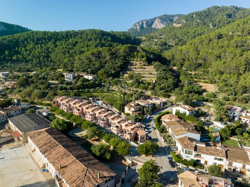 Drönarvy som visar moderna terrakottafärgade hus i en medelhavsby omgiven av skogsklädda berg.