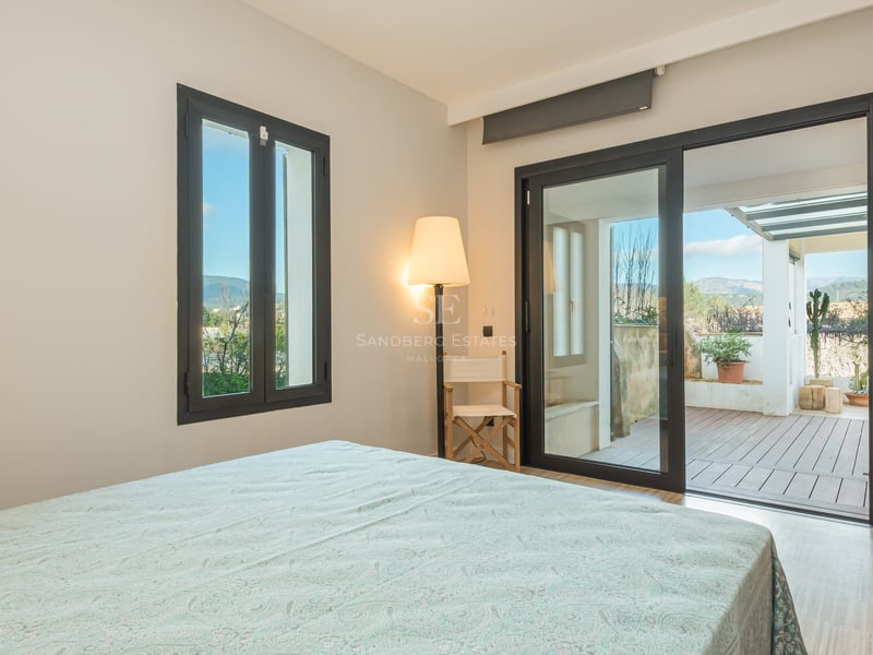 Ljust sovrum med skjutdörrar i glas som leder ut till en träterrass med bergsutsikt.