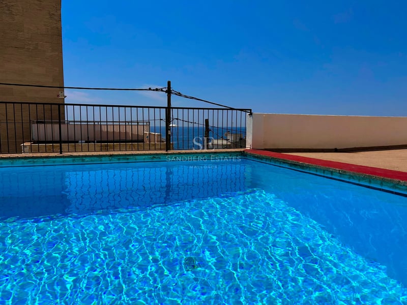Clear blue rooftop swimming pool with a black railing overlooking the sea under a bright blue sky.