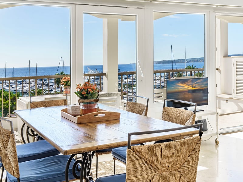 Sunlit dining room with a wooden table and woven chairs looking out over a yacht-filled marina and the blue ocean.