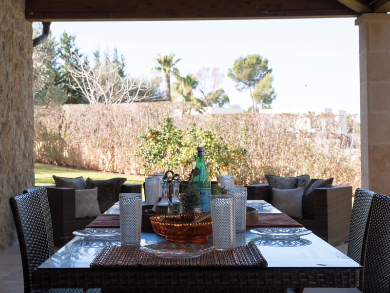 Övertäckt medelhavsterrass med matgrupp i konstrotting, naturstensväggar och utsikt över en lummig trädgård.