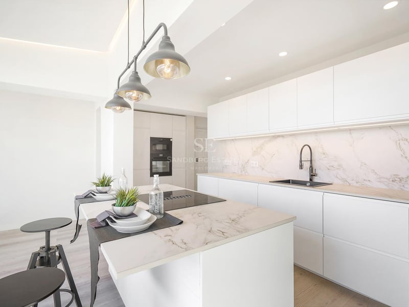 Moderne weiße Küche mit großer Marmorinsel, integrierten Geräten und industriellen Pendelleuchten.