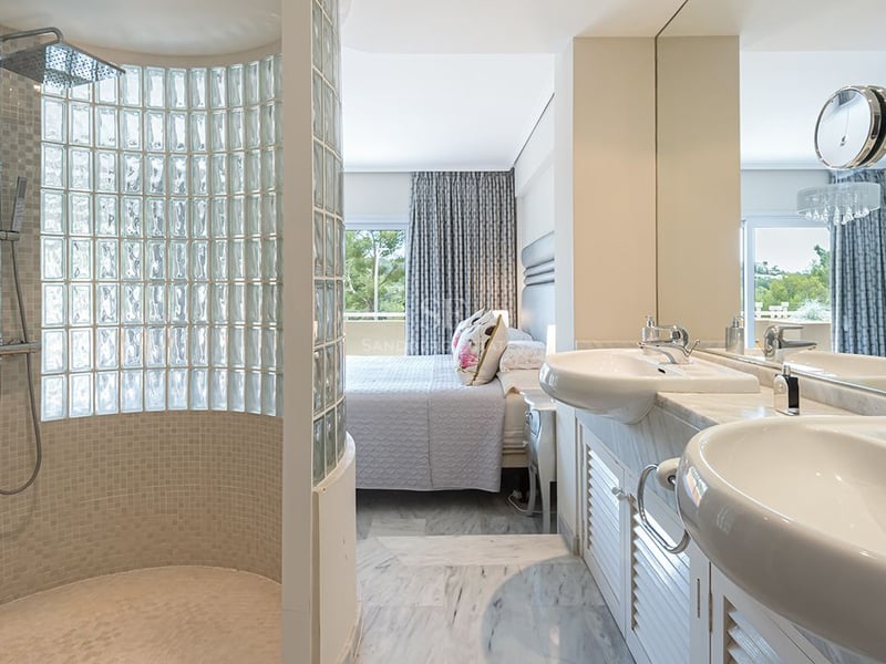 En-suite bathroom featuring double marble vanity and glass block shower, adjacent to a bright bedroom area.