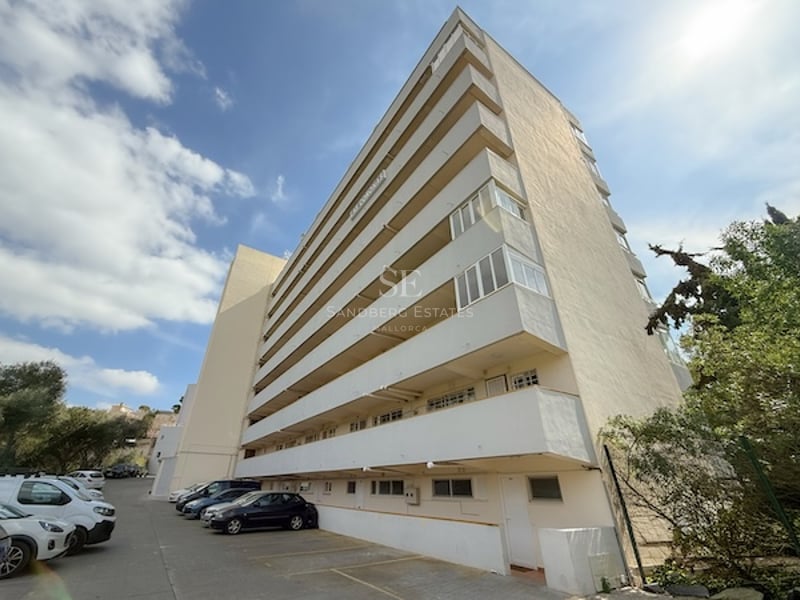 Vista exterior de un edificio de apartamentos blanco de varias plantas con amplios balcones y zona de aparcamiento.