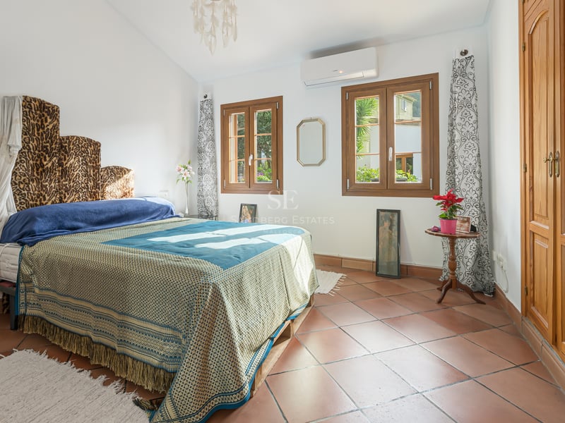 Sunny bedroom with terracotta tile floors, large wooden built-in wardrobes, and windows overlooking greenery.