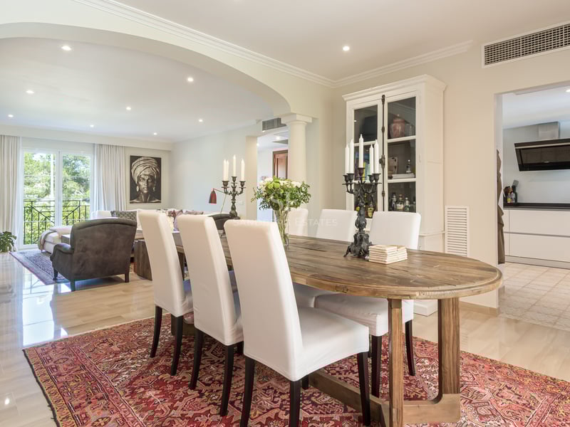 A rustic wooden dining table with white chairs on a red patterned rug, overlooking a living area and modern kitchen.