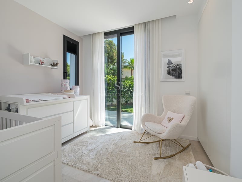 Chambre de bébé minimaliste avec berceau blanc, table à langer, fauteuil à bascule en bouclé et baie vitrée sur jardin.