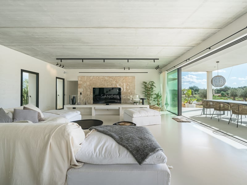 Modernt vardagsrum med vita soffor, betongtak, stenvägg och skjutdörrar i glas som leder ut till en terrass.