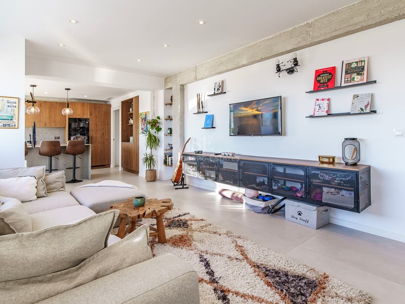 Grand salon avec canapé d'angle beige, tapis moelleux, poutres en béton brut et cuisine ouverte en arrière-plan.