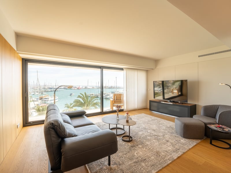 Modernt vardagsrum med lädersoffa och stora glaspartier med utsikt över en hamn med båtar.