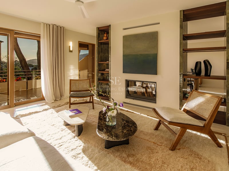 Helles Wohnzimmer mit Rattanstühlen, Marmortischen, Einbaukamin und großen Glastüren mit Blick ins Freie.