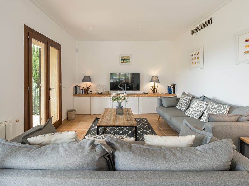 Salon lumineux avec canapé d'angle gris, table basse en bois et portes-fenêtres ouvrant sur le jardin.
