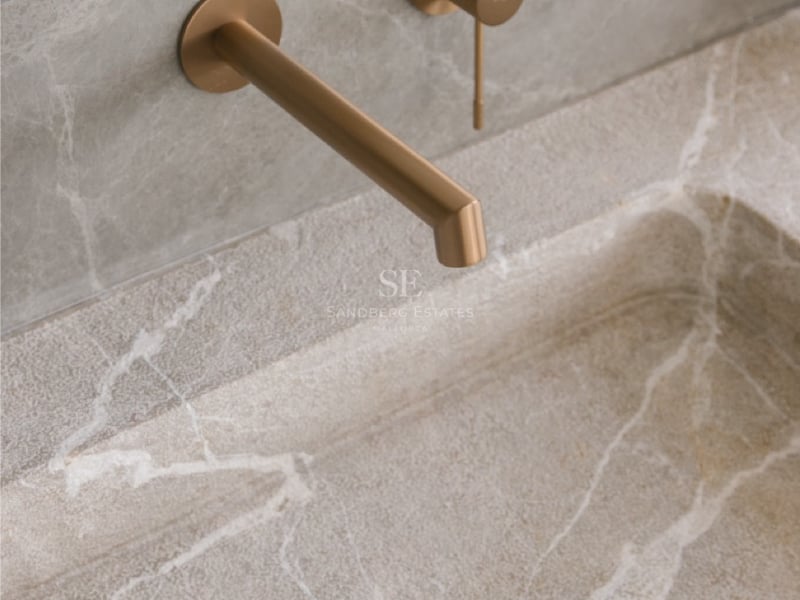 Wandmontierte Armatur aus gebürsteter Bronze über einem Waschbecken aus Naturstein mit weißer Maserung.