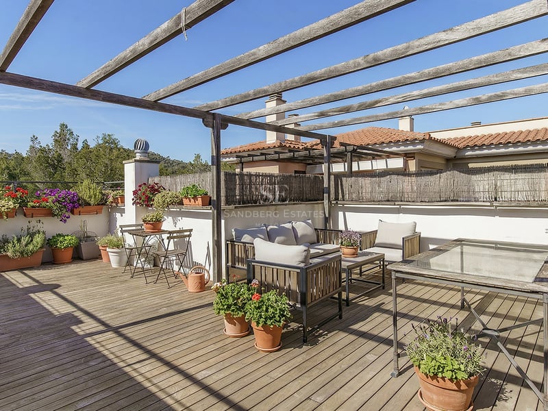 Takterrass med träpergola, trädäck, olika krukväxter och utemöbler under en klarblå himmel.