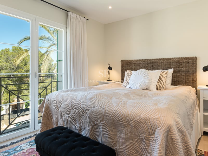 Bright bedroom featuring a woven wicker headboard, beige quilted bedding, and large French doors overlooking palm trees.