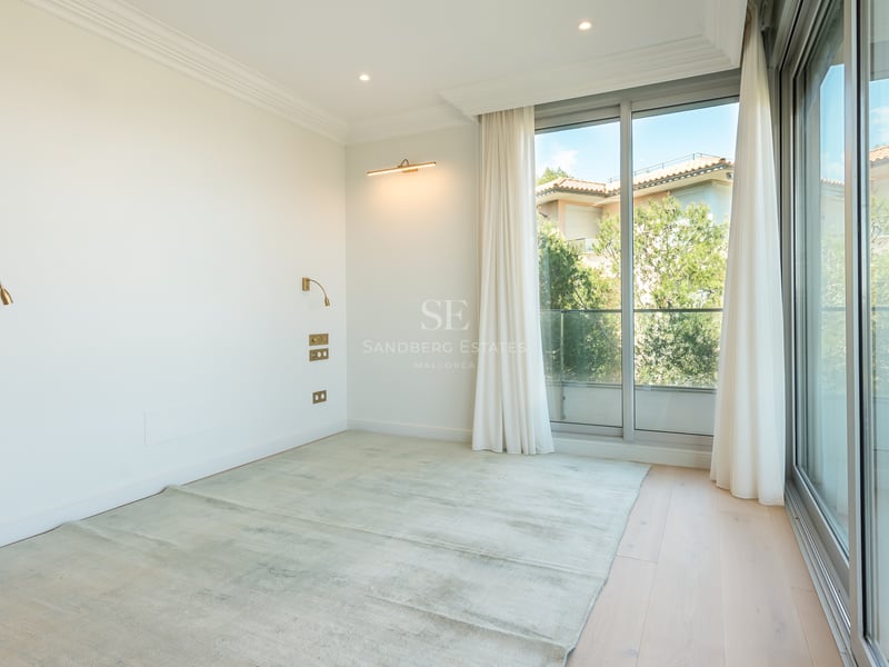 Empty modern bedroom with light wood floors, floor-to-ceiling glass doors, white walls, and elegant brass wall lamps.