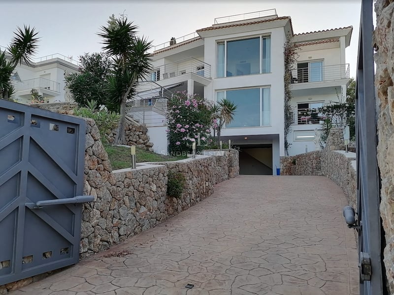 Villa blanche à plusieurs niveaux avec grandes baies vitrées, vue depuis une allée sécurisée avec murs en pierre.