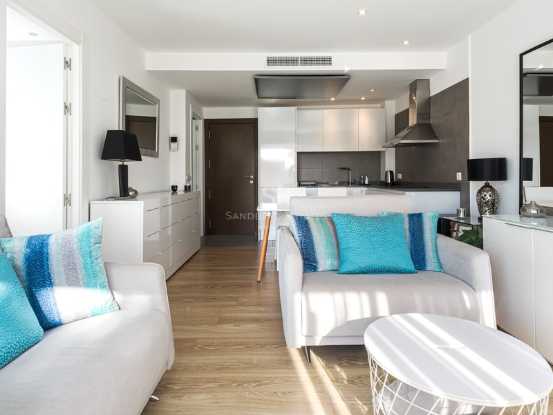 Salon lumineux avec canapés blancs, coussins turquoise, parquet en bois et cuisine moderne en arrière-plan.