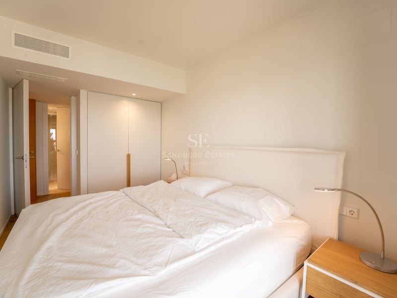 Ljust sovrum med stor säng, vita sängkläder, inbyggd garderob och sängbord i trä.