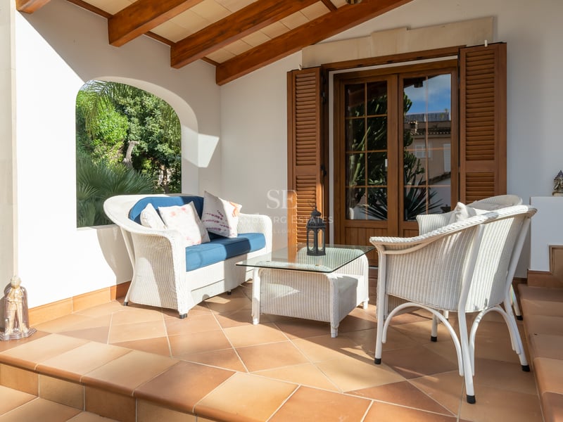 White wicker furniture on a terracotta-tiled terrace with exposed wooden beams and an arched view of a garden.